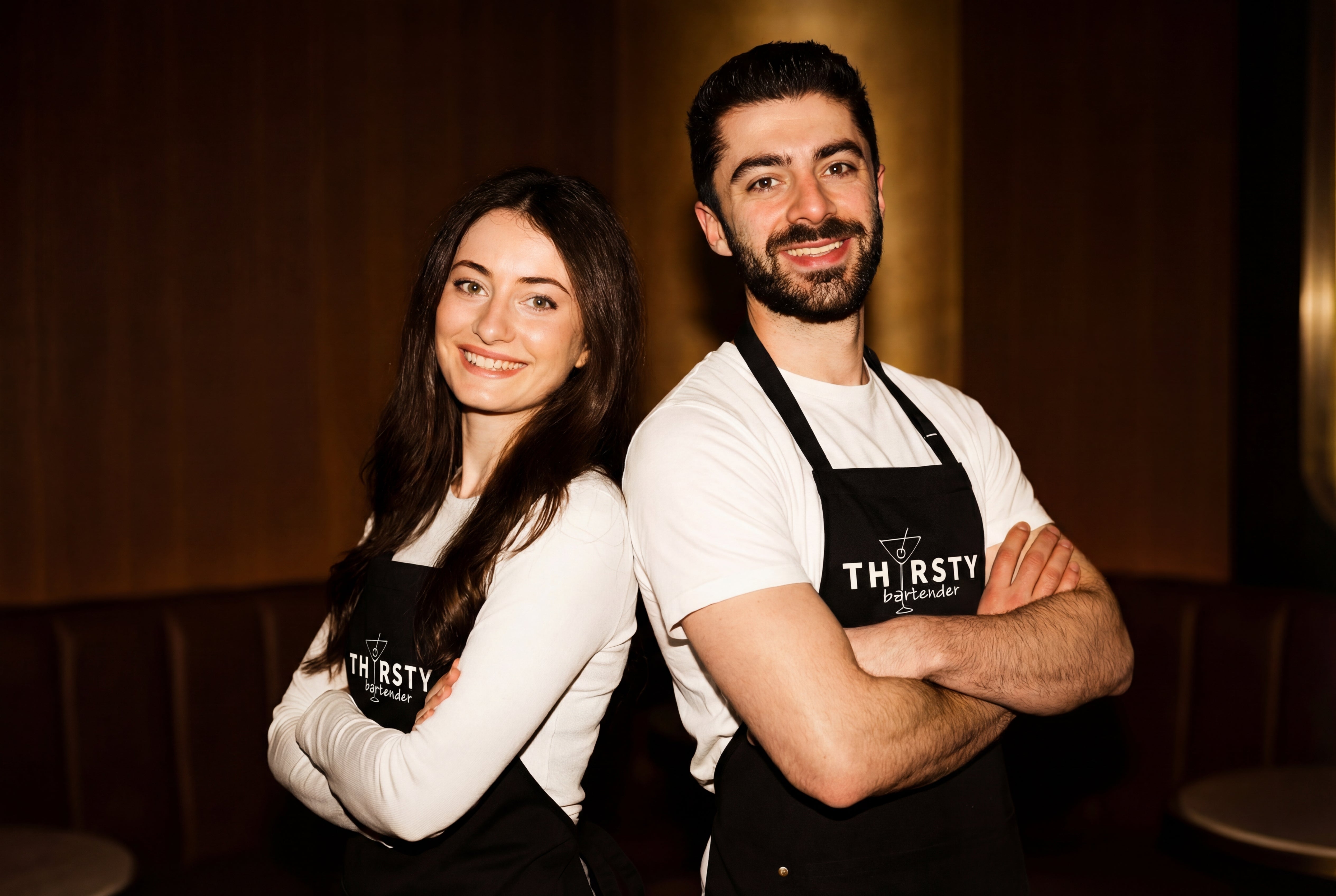 Two people wearing aprons with 'Thirsty Bartender' logo in a dark background setting
