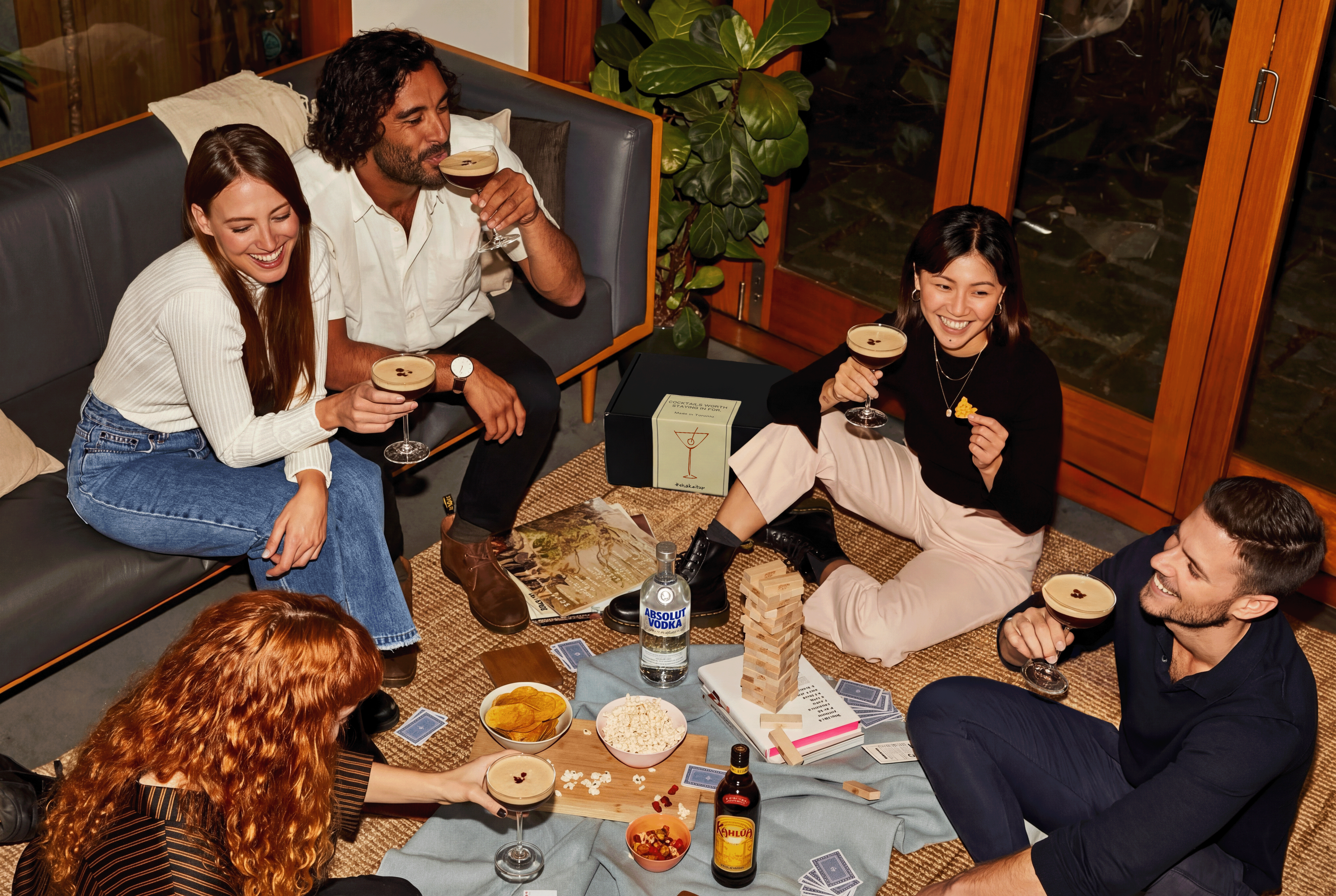 Group of people sitting on a couch and floor, enjoying drinks and snacks in a casual setting.