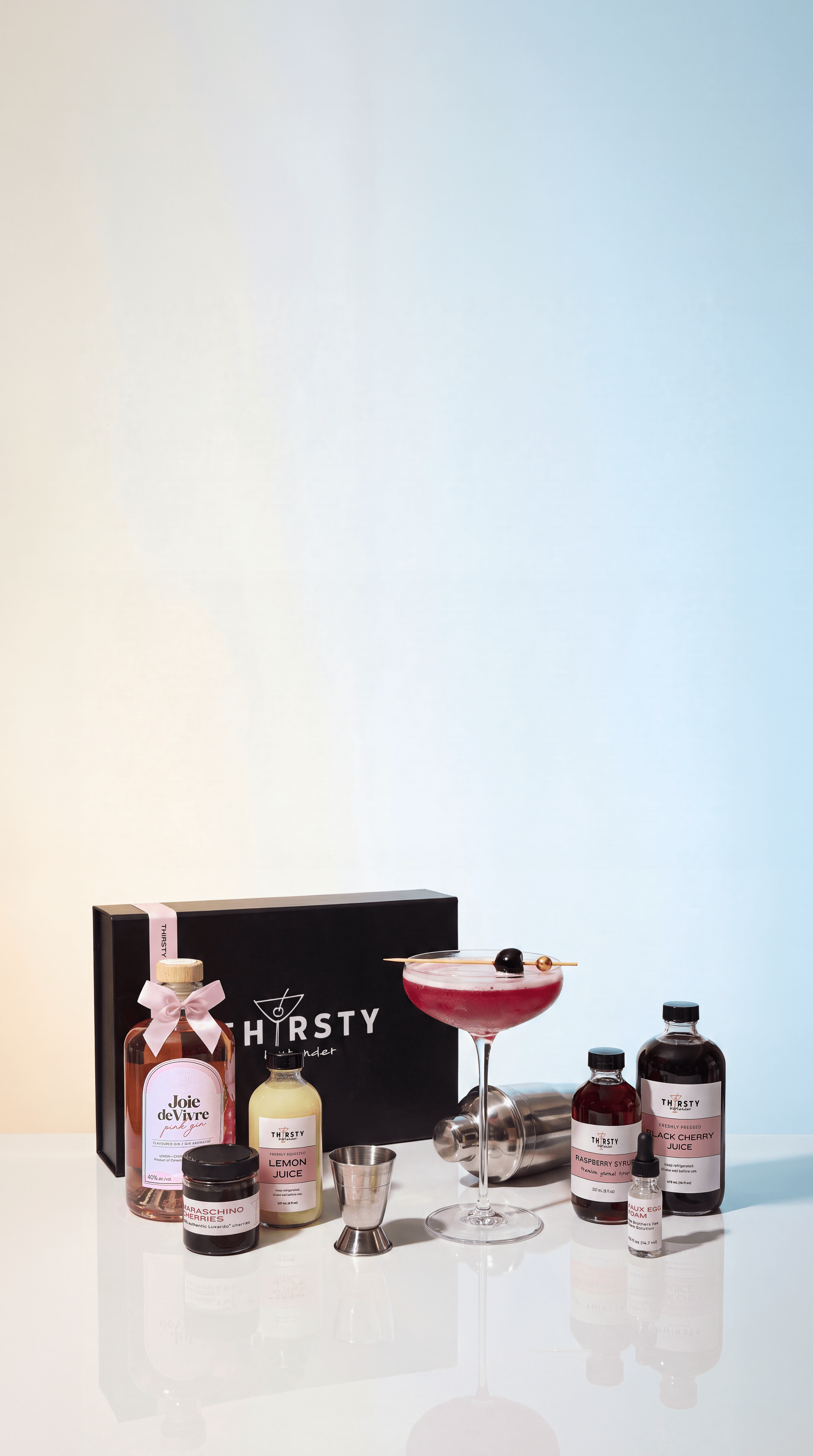 A Tirsty Bartender cocktail kit arranged on a reflective white surface against a light blue gradient background.