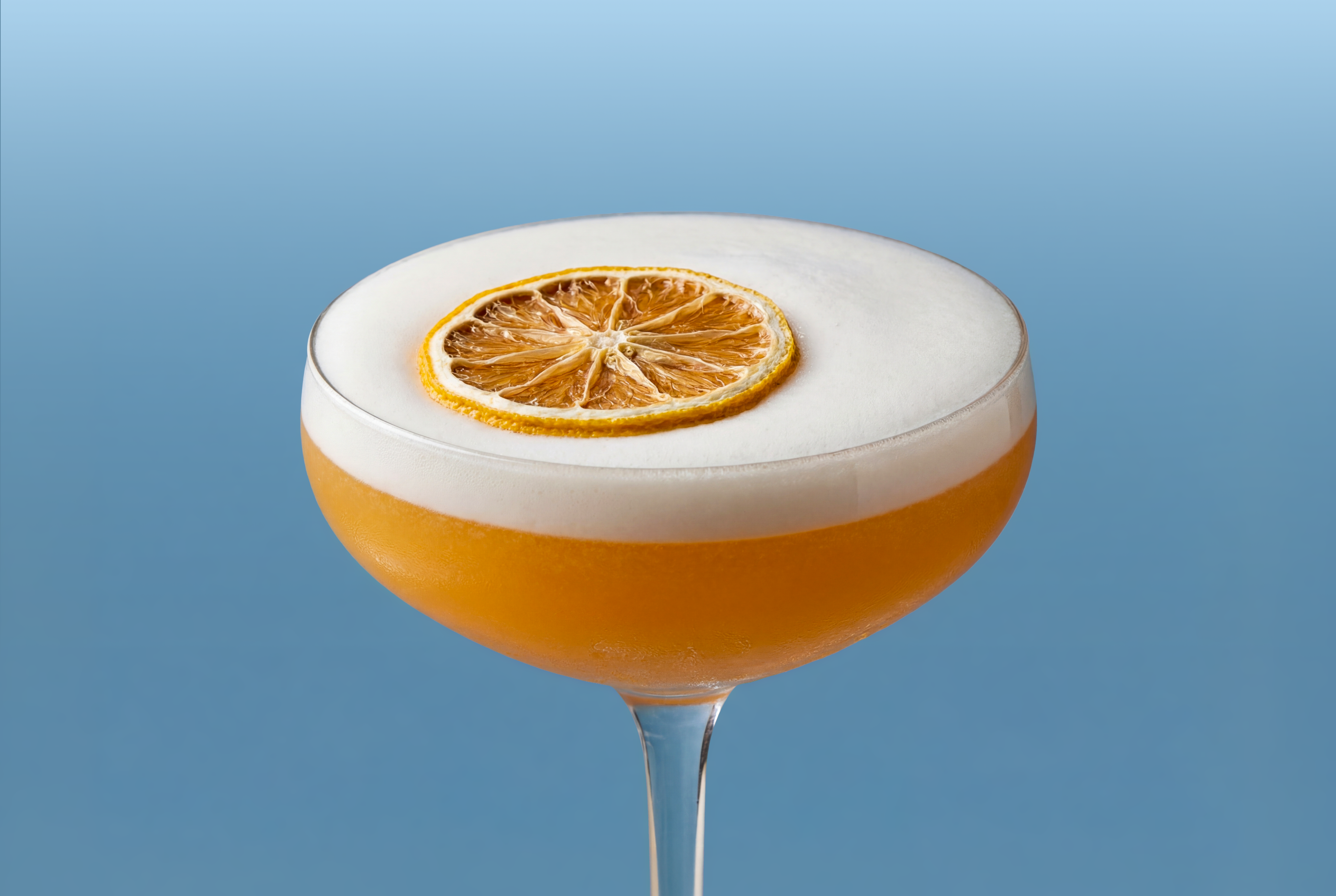 Whiskey sour cocktail glass with a white foam top and orange slice garnish on a blue background